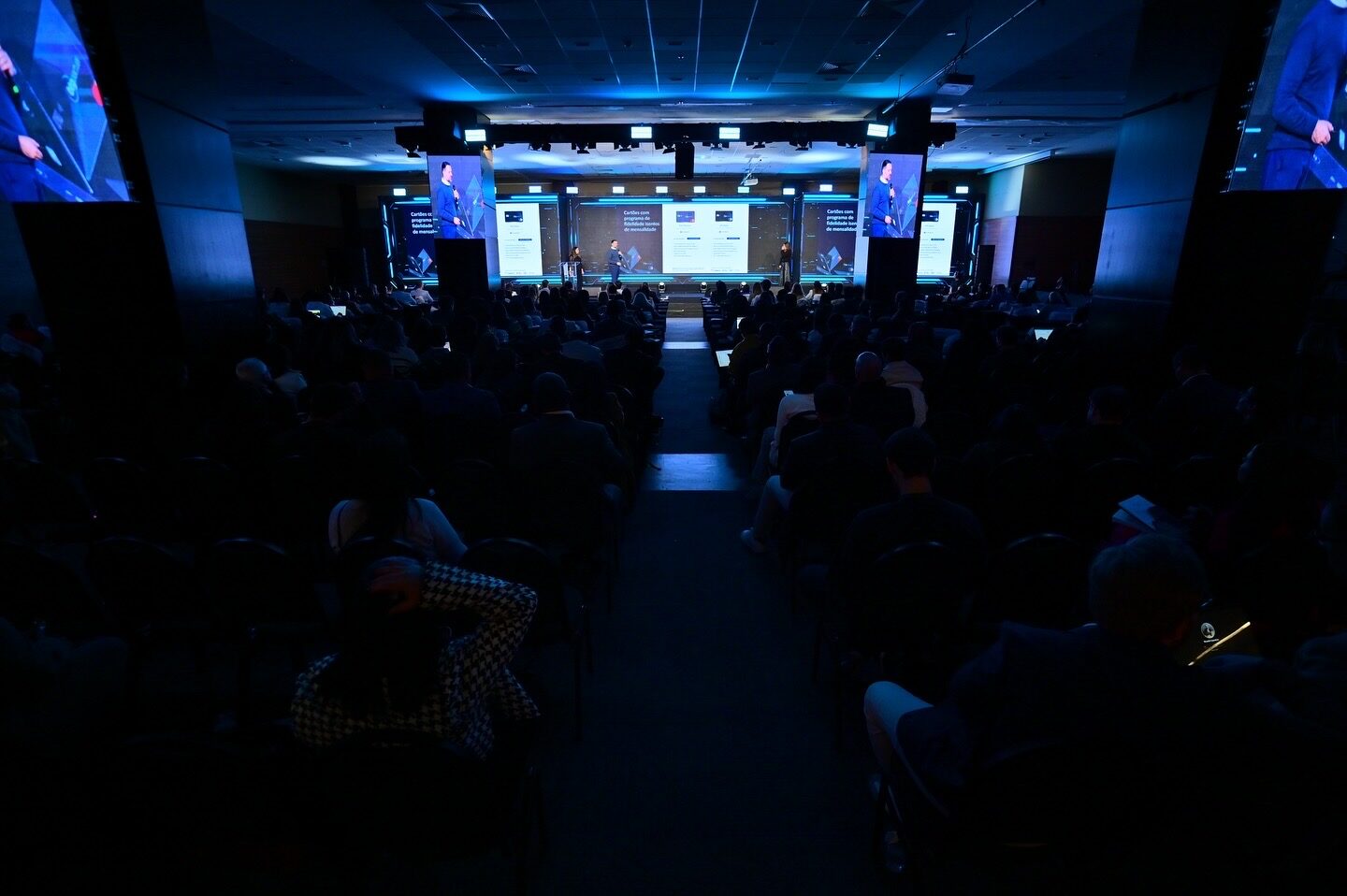Auditório lotado em salão de hotel com painéis de LED azuis ao redor do palco corporativo