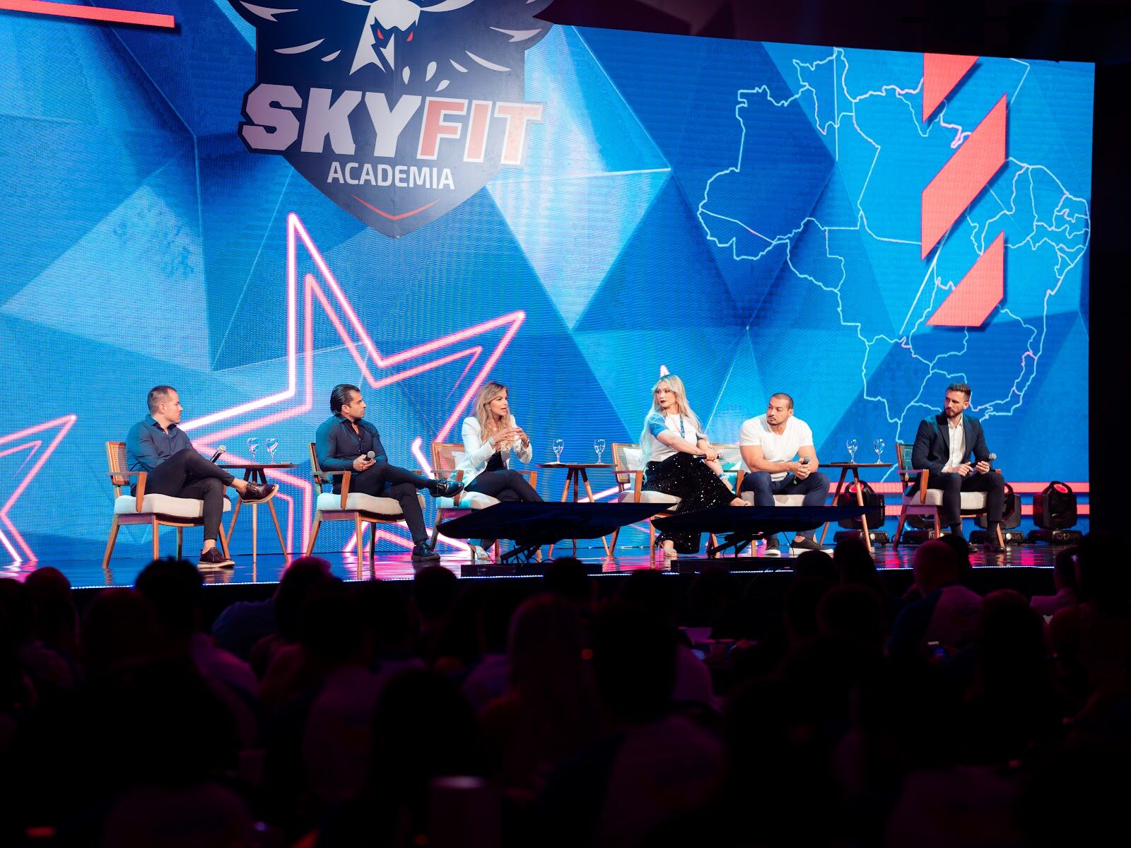 Participantes em mesa redonda no palco com grande painel de LED ao fundo em evento corporativo