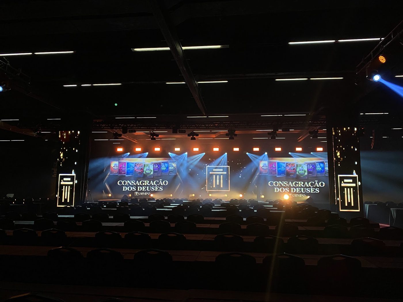 Palco de evento corporativo com feixes de luz laranja e azul e telões de LED ao fundo