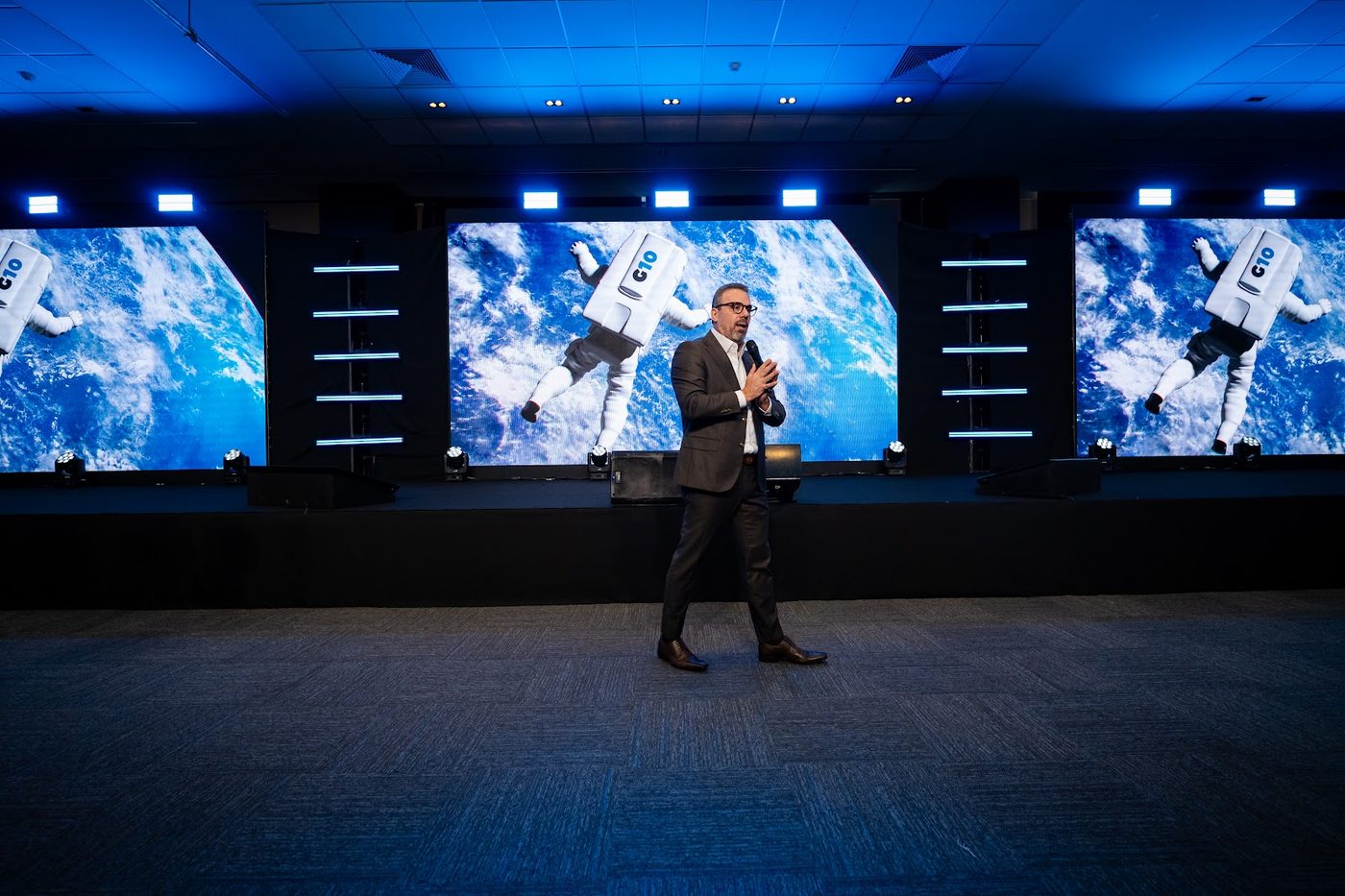 Palestrante em palco corporativo com três telões de LED ao fundo exibindo conteúdo visual do evento