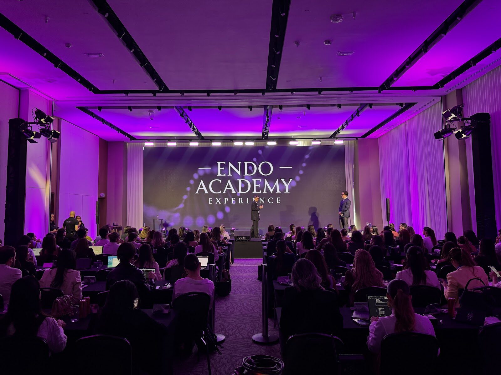Palco de evento corporativo com dois apresentadores e plateia sentada em mesas redondas