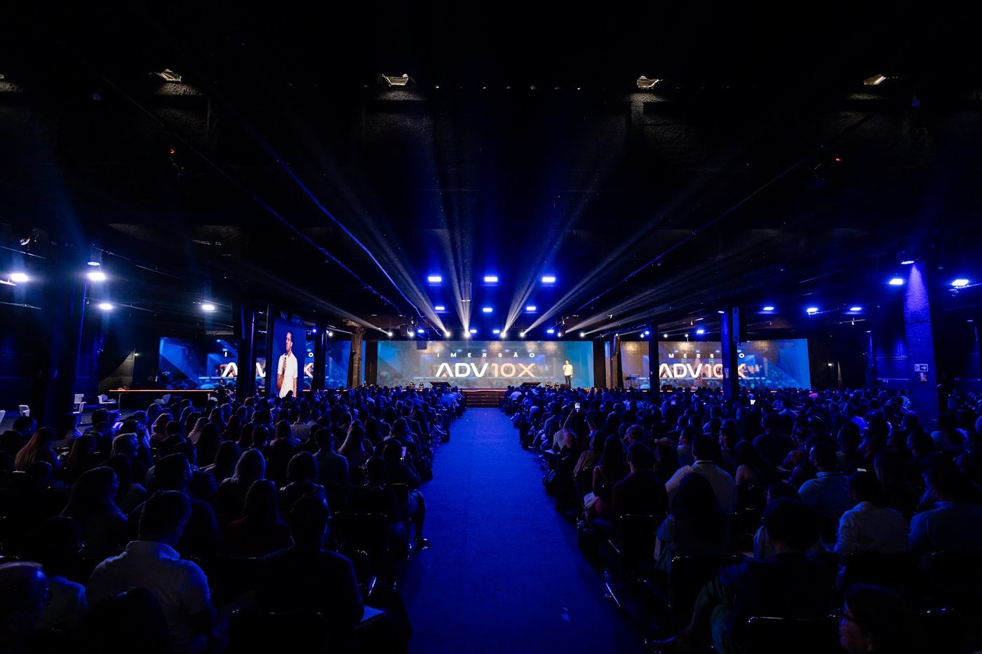 Vista panorâmica de auditório lotado com feixes de luz azul, painéis de LED e palco ao fundo