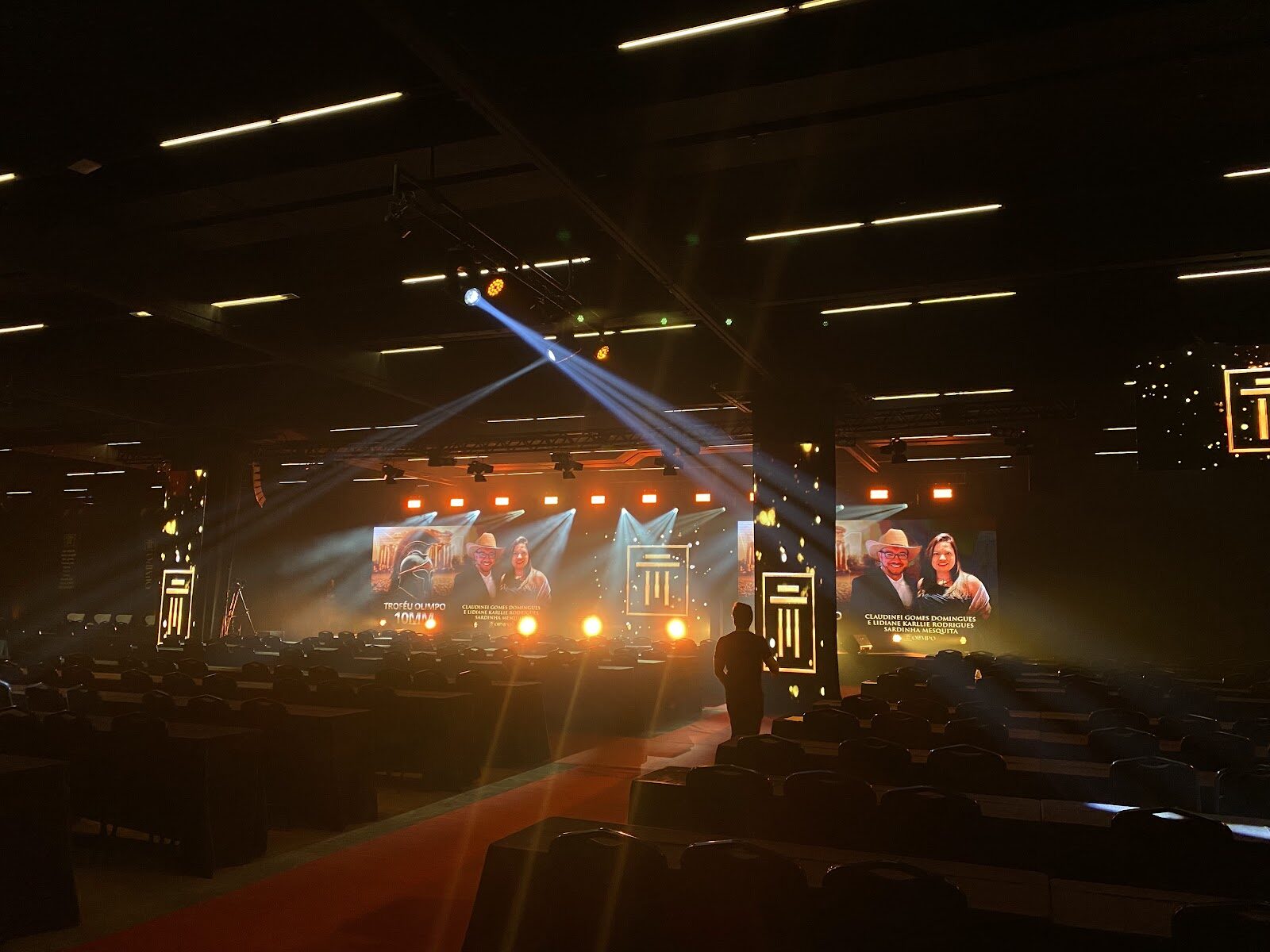 Palco de premiação corporativa com confete dourado, telões LED e feixes de luz azuis