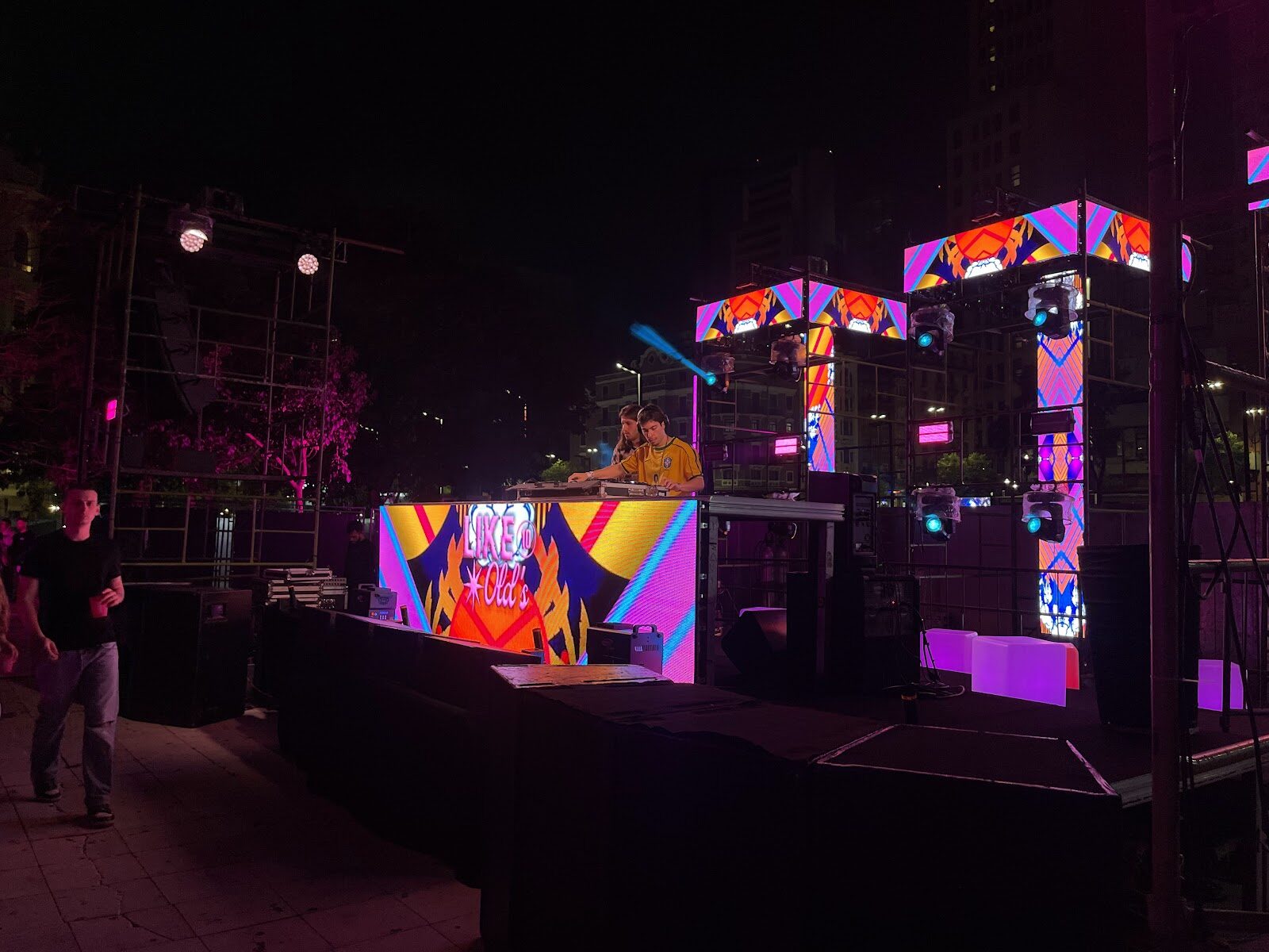 Palco com telões de LED e iluminação em evento ao ar livre à noite, skyline urbano ao fundo