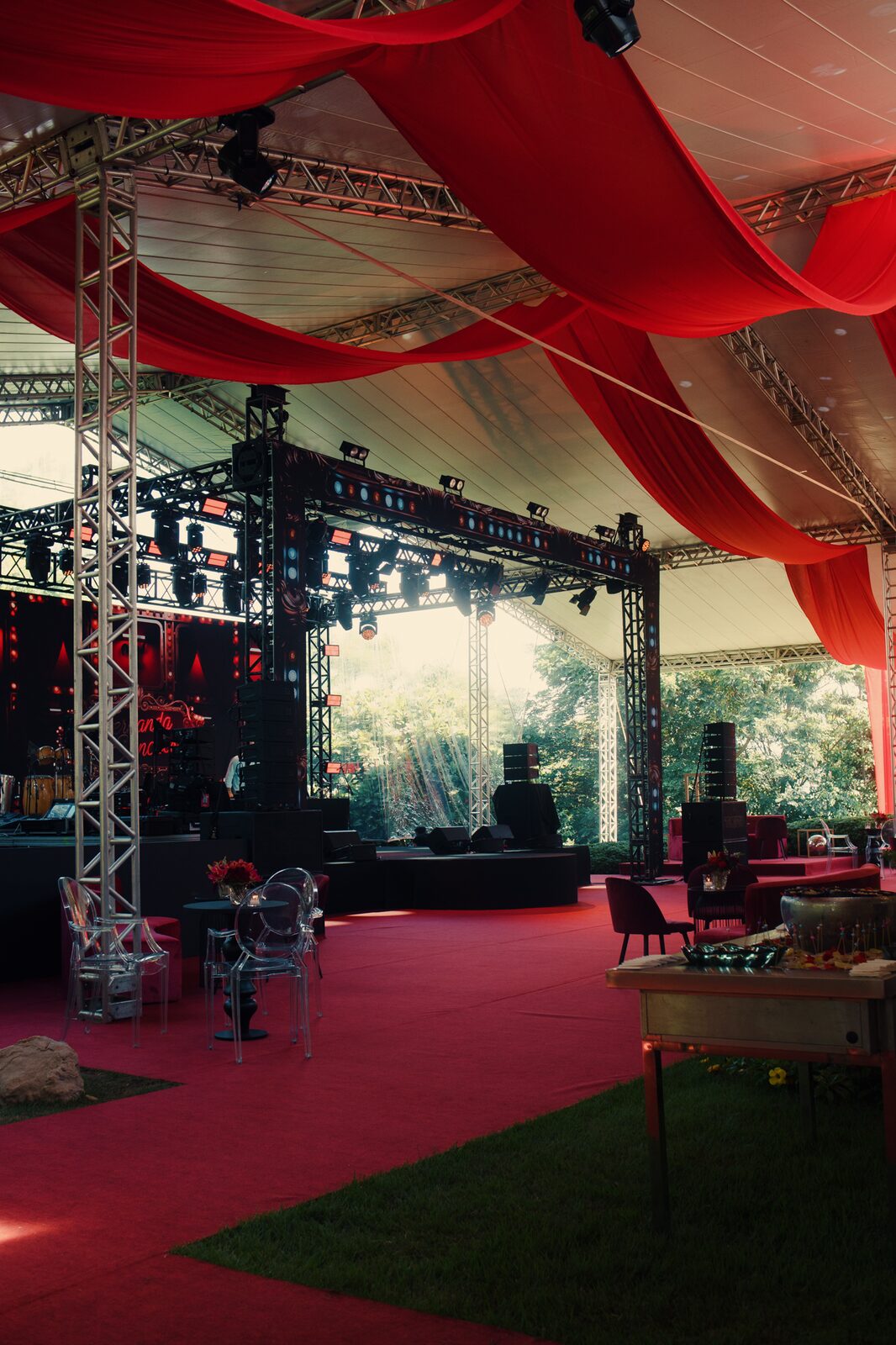 Palco com treliça e iluminação cênica montado em tenda ao ar livre, mesas de evento no primeiro plano
