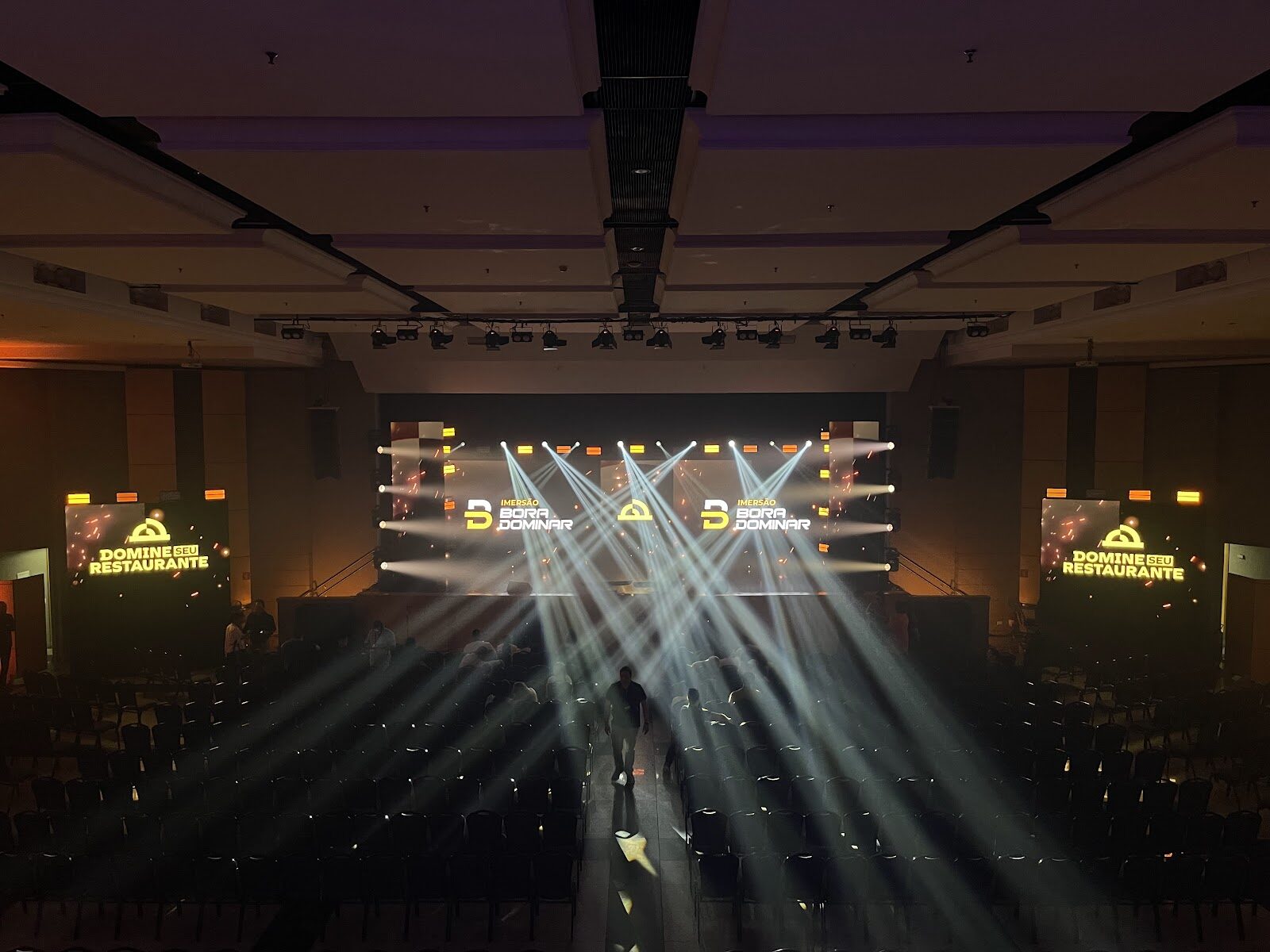 Auditório com feixes de luz cênica e telões laterais de LED exibindo conteúdo do evento