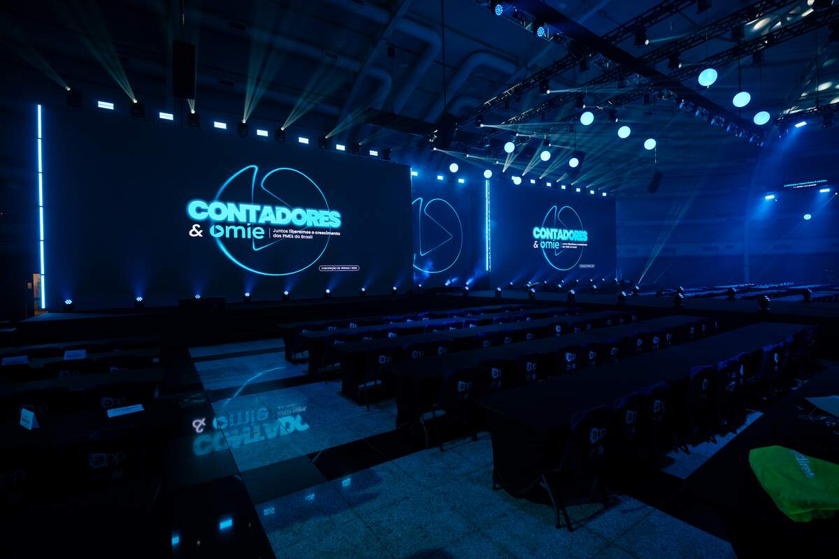Auditório com telões de LED em formato ultrawide exibindo identidade do evento, com iluminação cênica azul