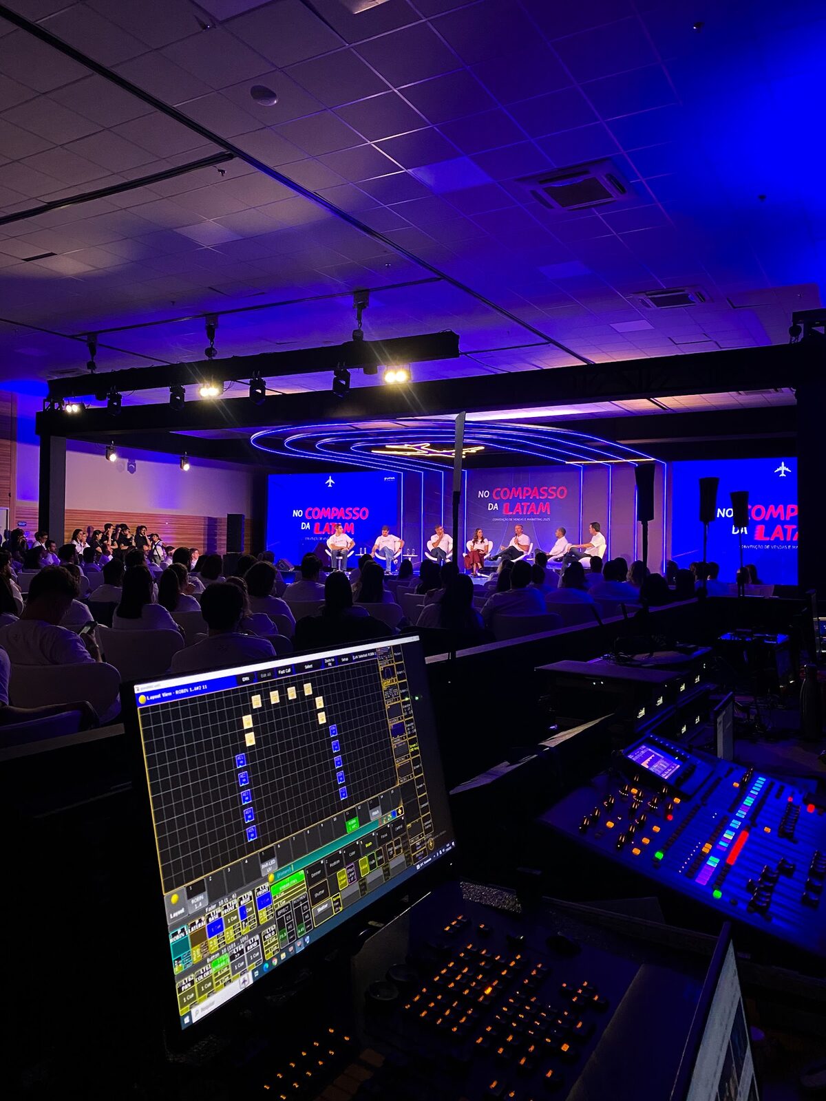Mesa de controle de iluminação com palco e telões LED ao fundo em evento corporativo