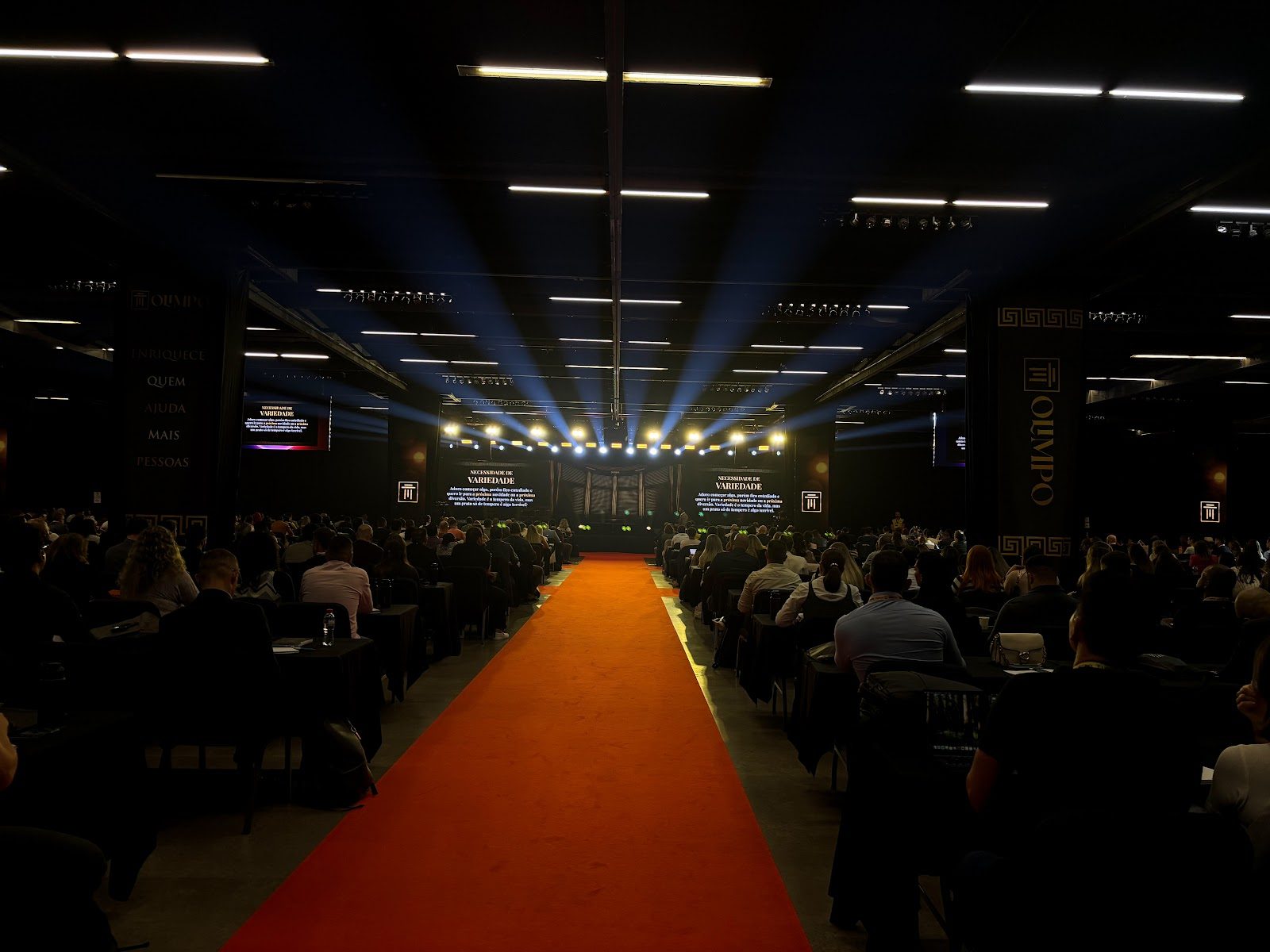 Feixes de luz convergindo no palco de convenção corporativa, com plateia lotada e tapete vermelho central