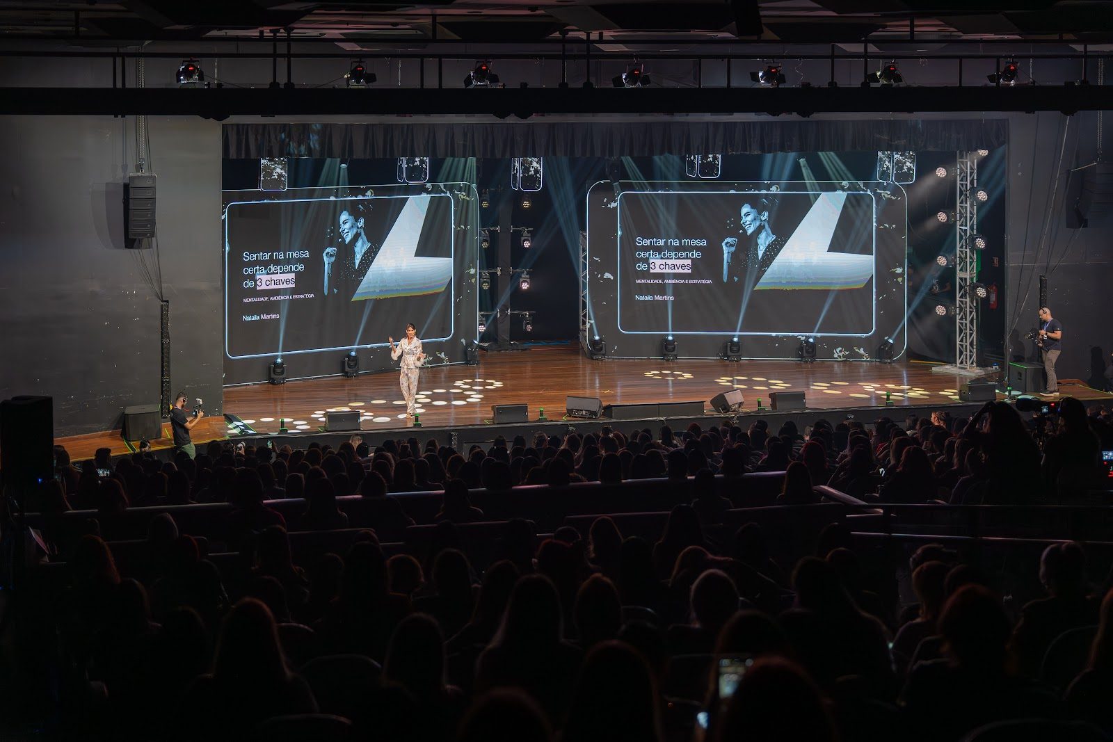 Palestrante iluminada pela luz frontal no palco corporativo, com moving heads na treliça e dois painéis de LED ao fundo