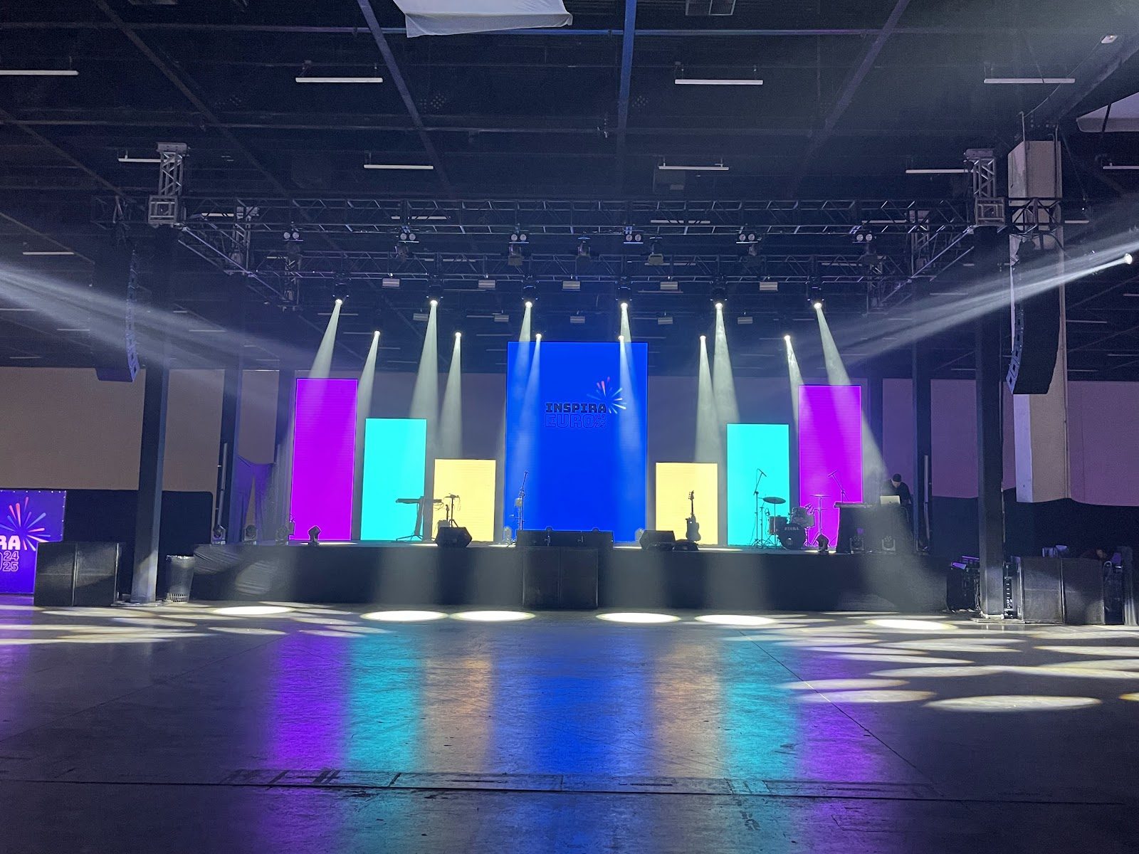 Grid de treliça com moving heads sobre palco corporativo, feixes de luz e gobos projetados no piso do pavilhão