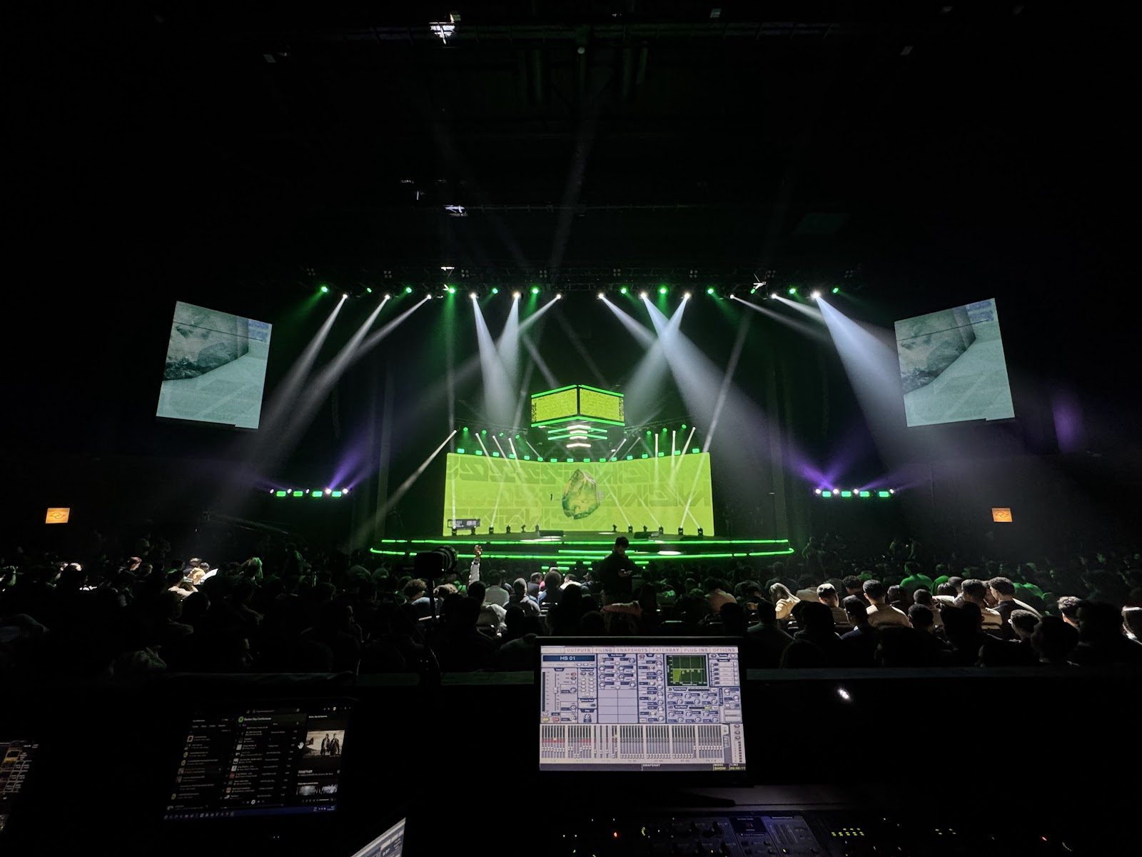 Mesa de operação de luz no FOH com tela do console aberta, palco com feixes verdes e plateia lotada ao fundo