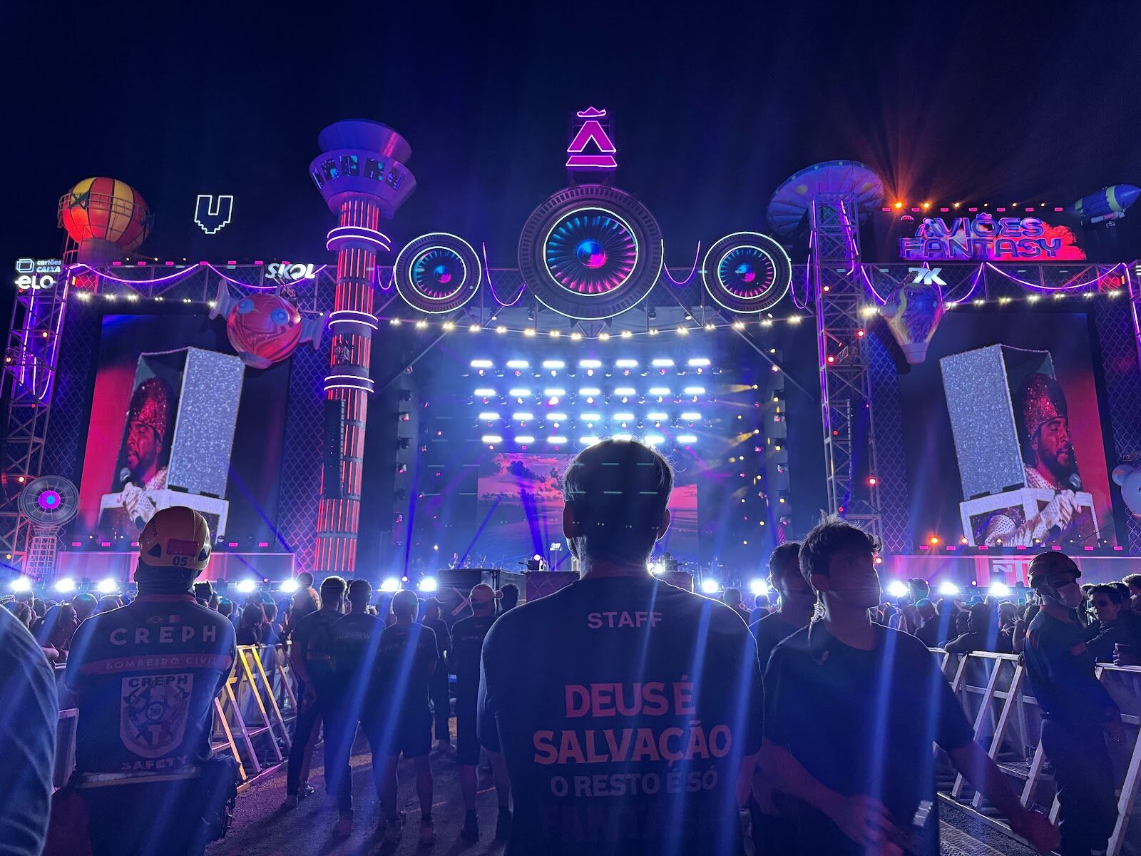 Festival com palco amplo, feixes de luz, painéis de LED e plateia vista de costas à noite