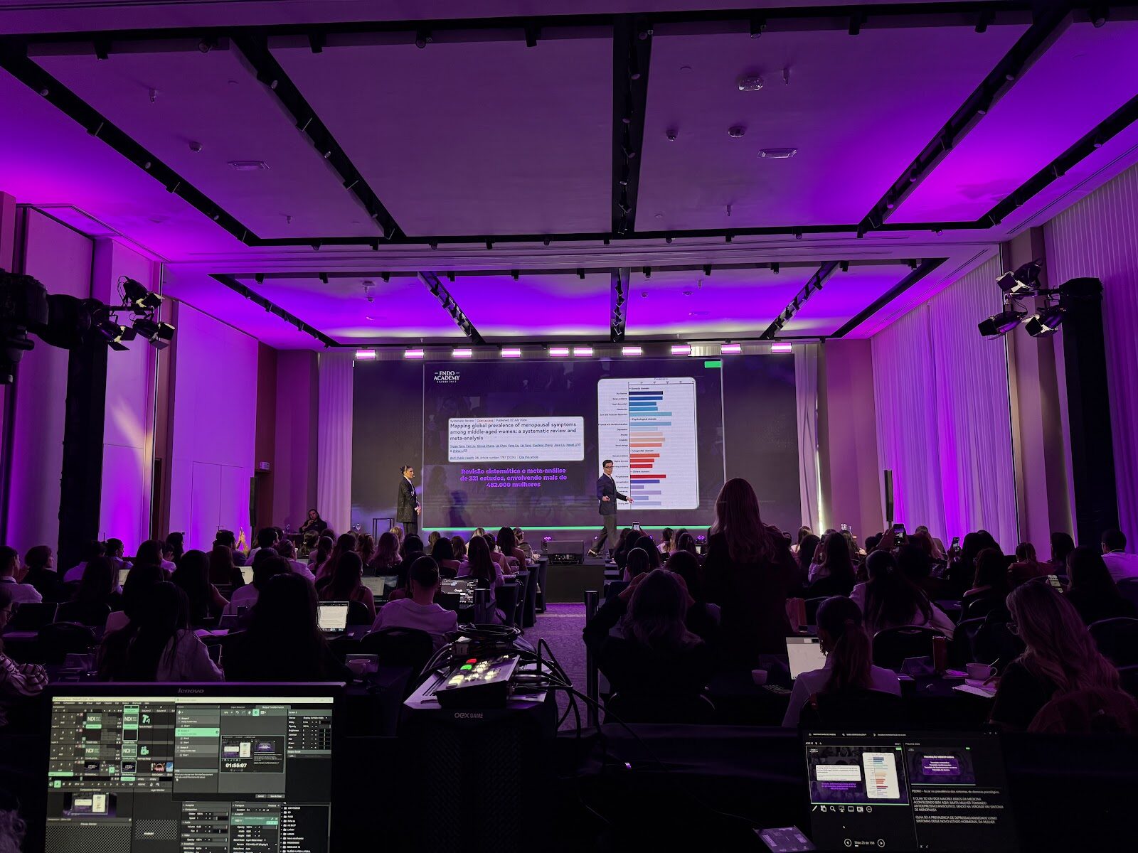 Mesa de operação técnica com monitores em primeiro plano e palestrante diante do painel de LED em evento corporativo