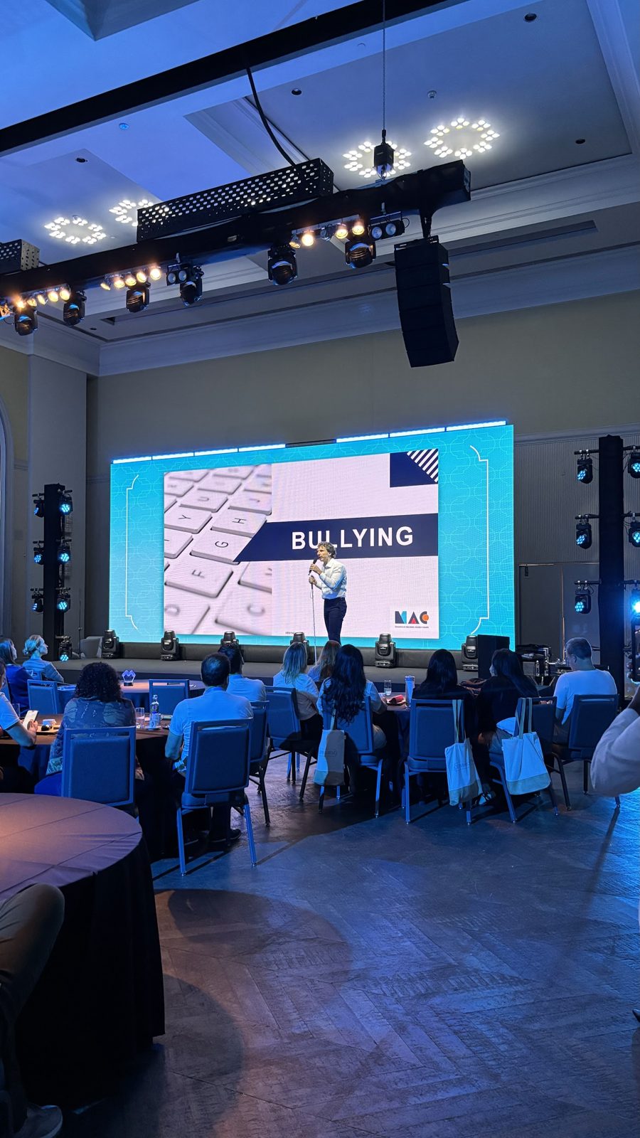 Line array suspenso ao lado do palco de evento corporativo com painel de LED, palestrante e plateia em mesas redondas