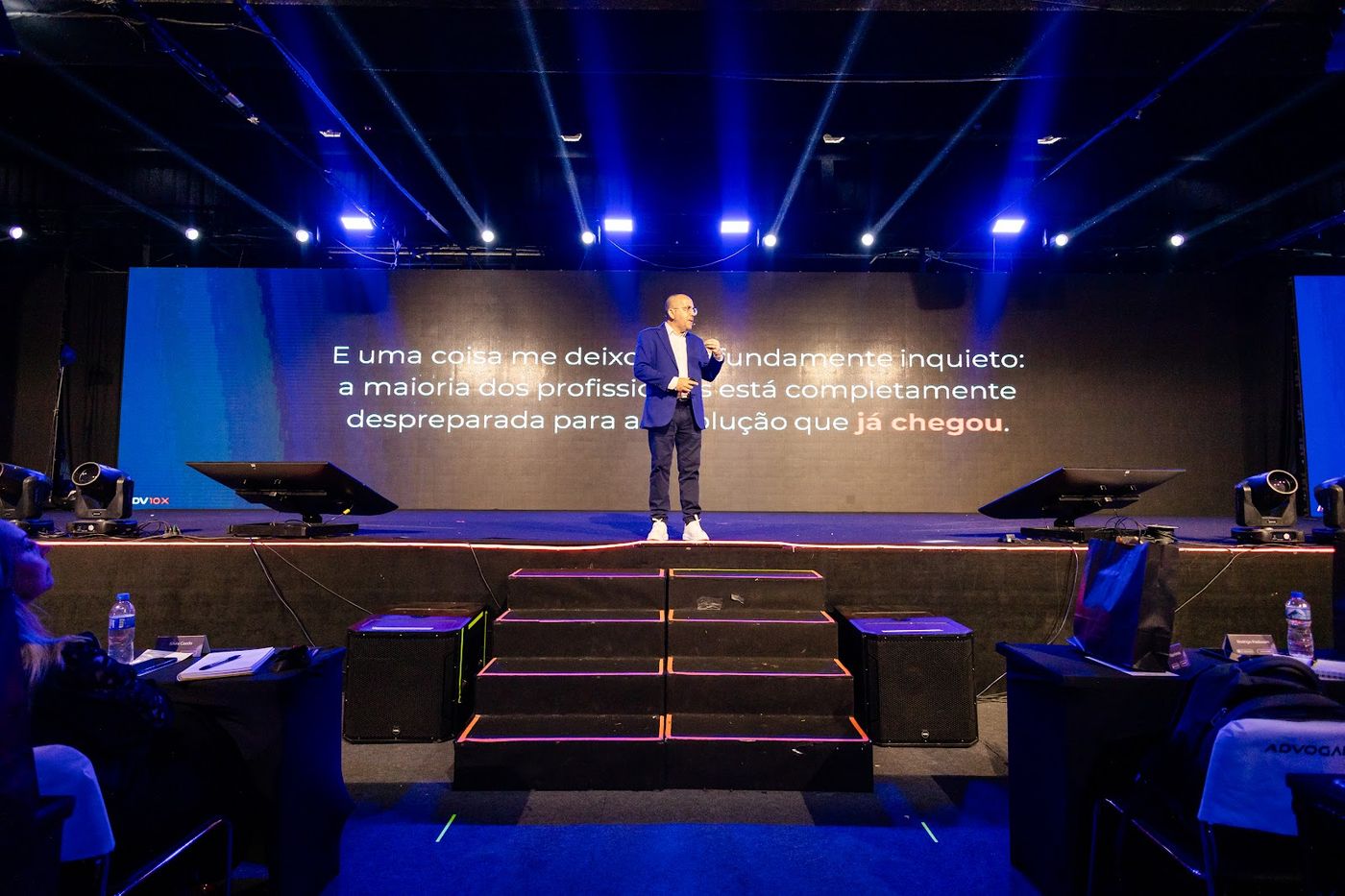 Palestrante em palco corporativo com caixas de som, telão LED e plateia ao fundo