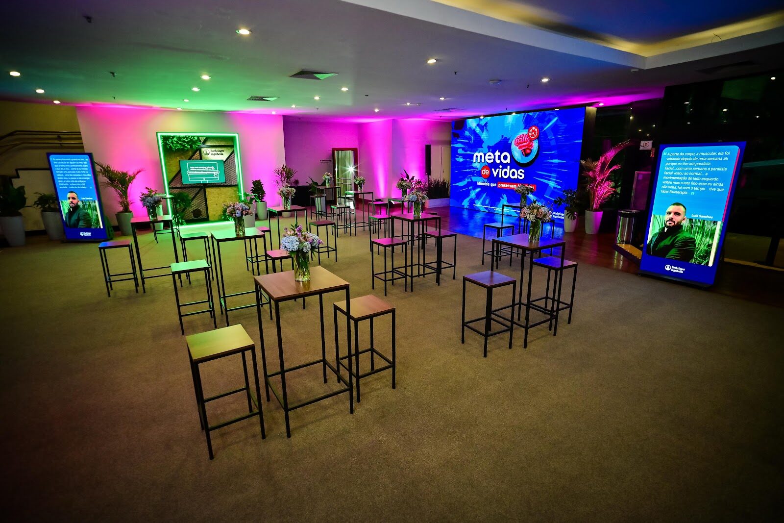 Lounge corporativo com painel LED colorido, mesas decoradas e iluminação cênica rosa e verde