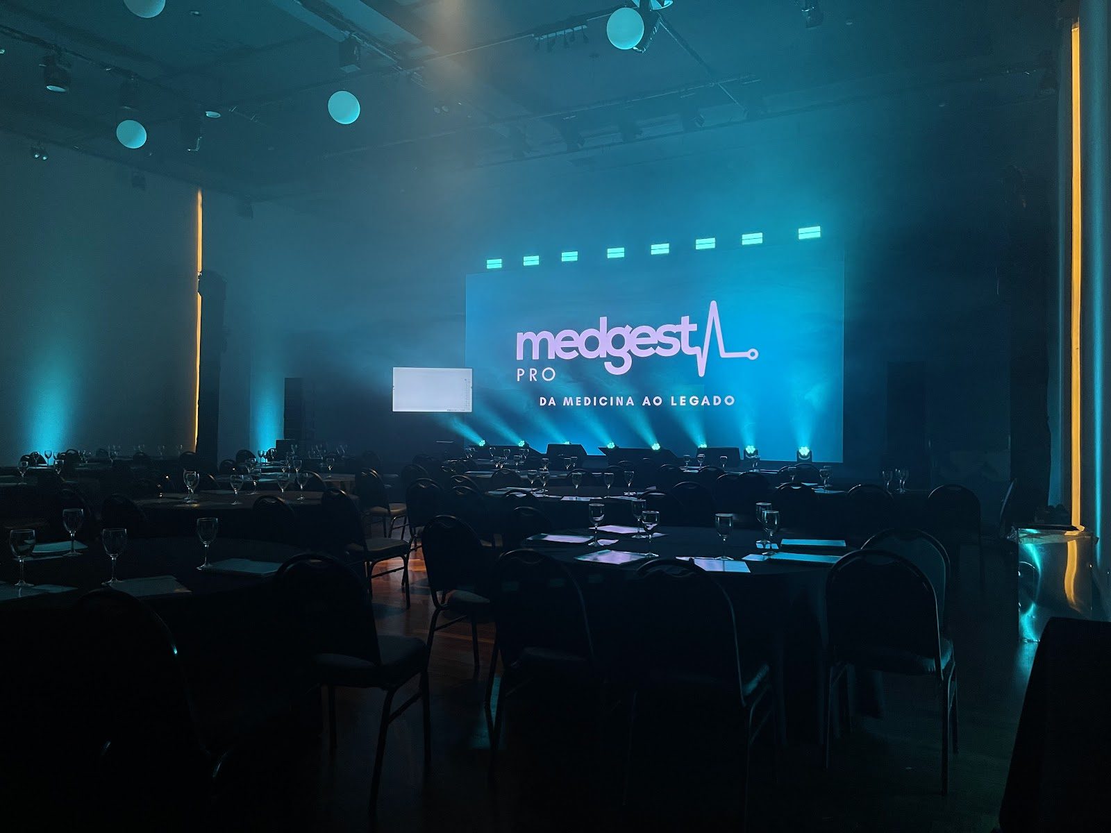 Salão de evento corporativo com uplighting azul e colunas de luz amarela nas laterais