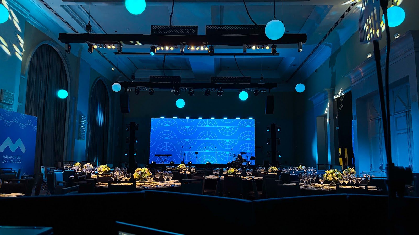 Salão corporativo com painel LED azul, esferas luminosas e mesas postas para jantar