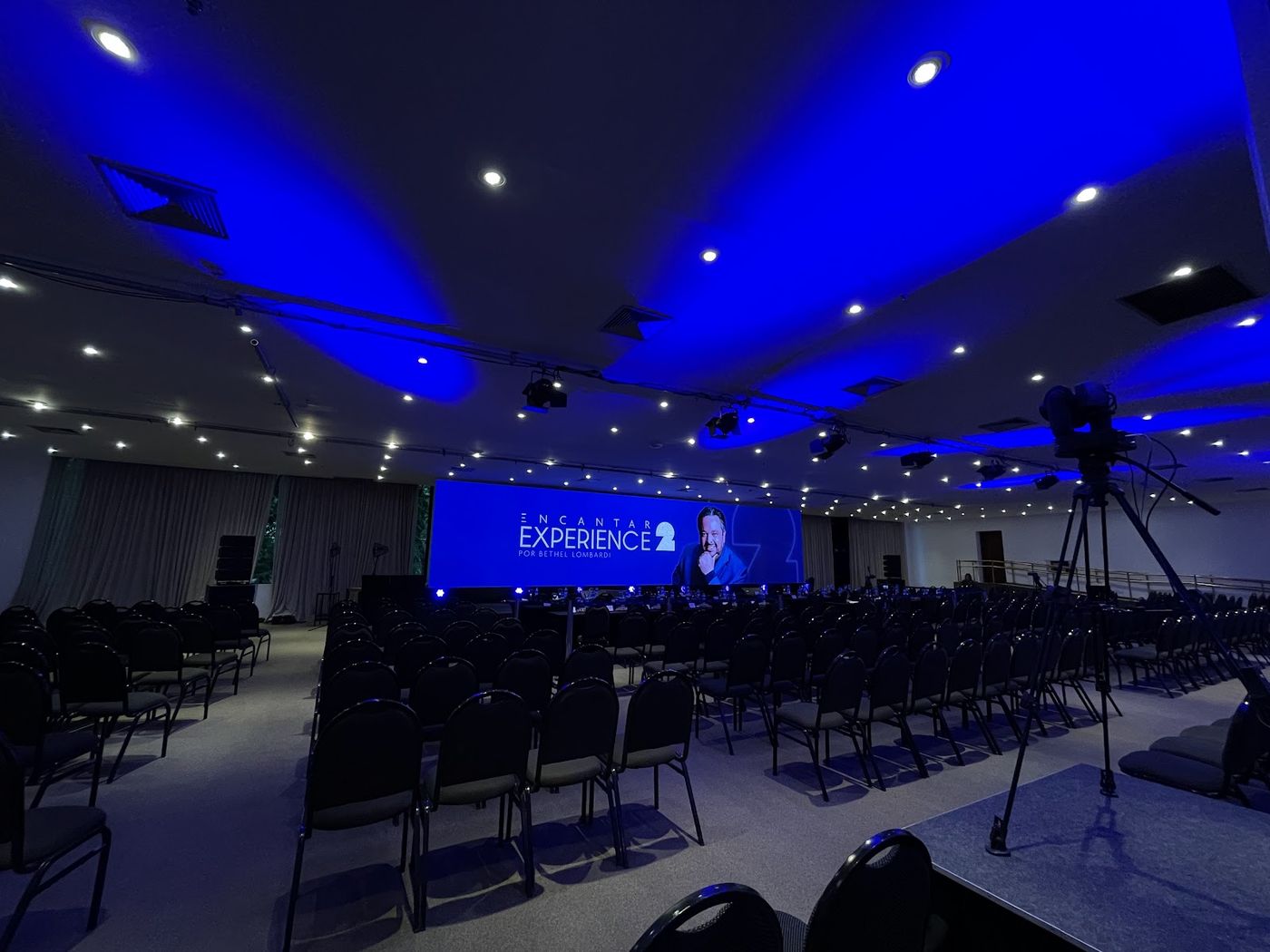 Auditório vazio com fileiras de cadeiras e câmera de transmissão diante de painel LED